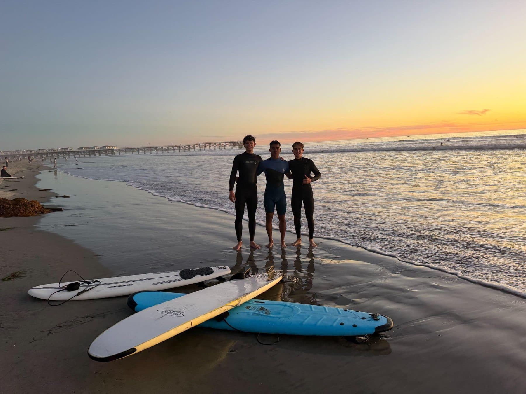 Three friends in wetsuits at the shoreline at sunrise with their boards on the sand